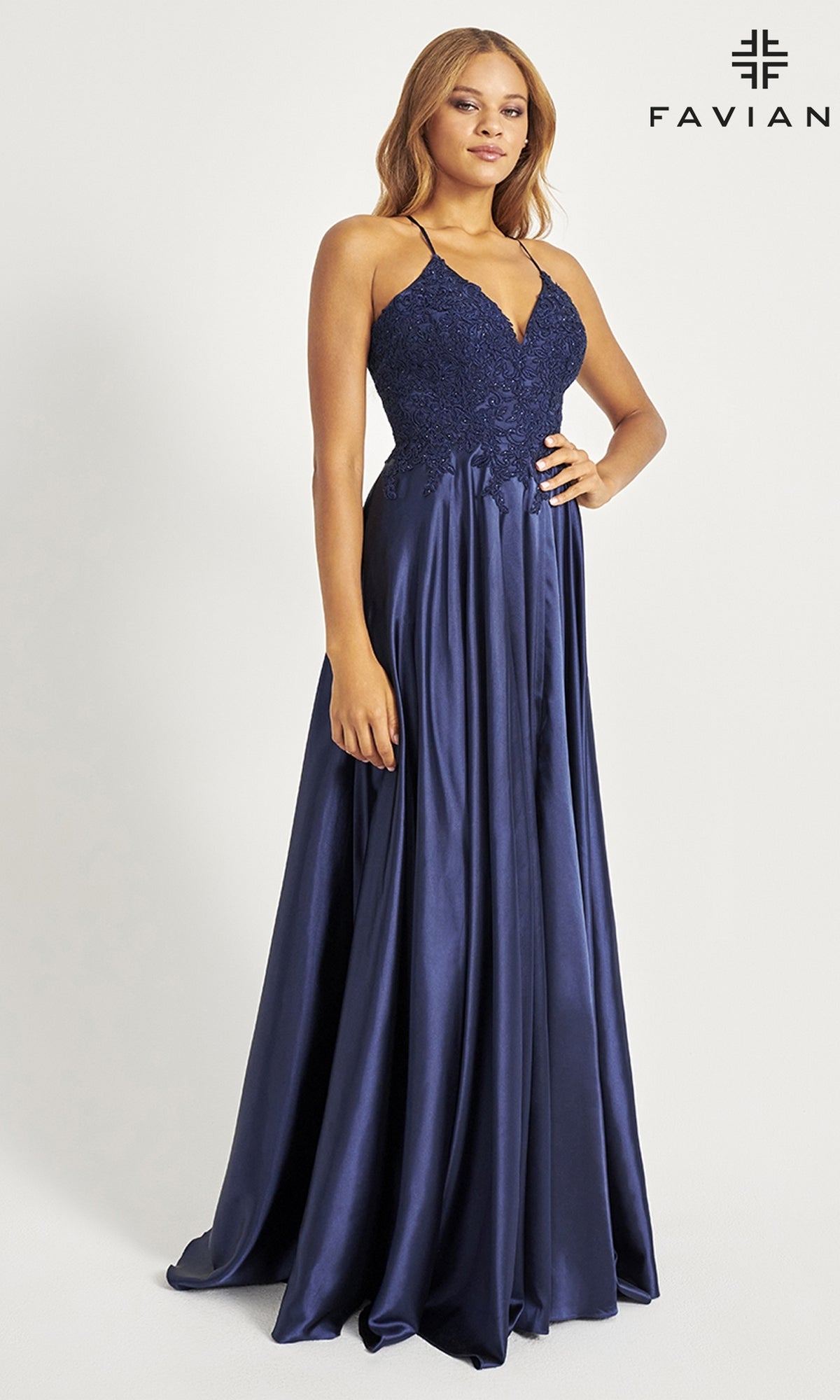 A woman wears the Faviana Lace-Bodice Long A-Line Prom Dress, featuring a navy blue embroidered lace bodice with thin straps and a flowing satin skirt. The background is plain and the Faviana logo is visible.